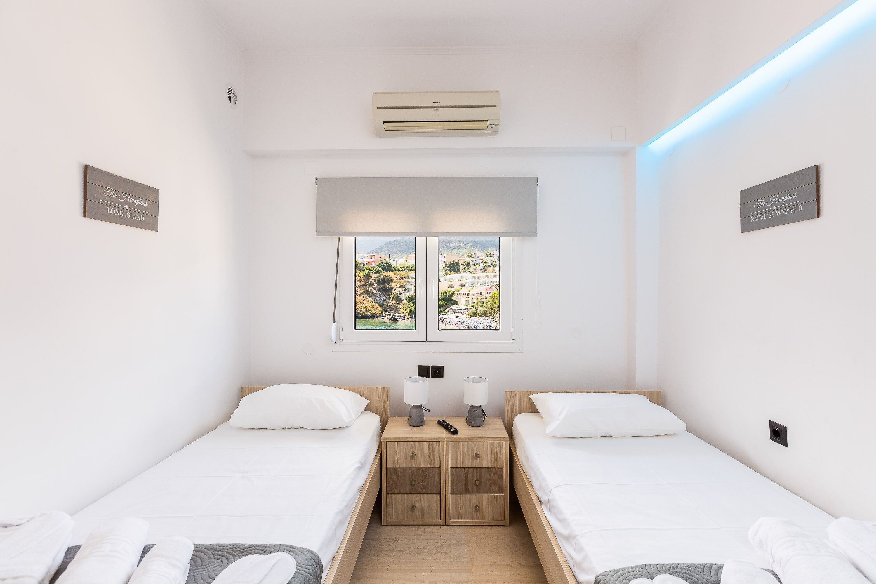 A small, clean room with two beds, a window with a view, an air conditioner, and a bedside table with a lamp.