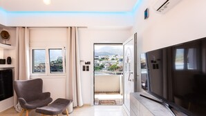A modern living room with a flat-screen TV, a grey armchair, a matching ottoman, a small table, and a view of a cityscape through a glass door. - Ocean Dream Suite (Mylopotamos)