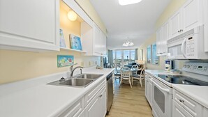 A modern kitchen with white cabinetry, a dishwasher, a microwave, and a dining area with a table and chairs. - Cozy Gulf Front Condo | Private Beach Access | Pool | Sleeps 8 (Gulf Shores)