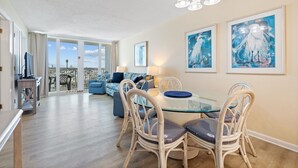 A spacious living area with a glass dining table and wicker chairs, a sofa, and a view through large windows. - Cozy Gulf Front Condo | Private Beach Access | Pool | Sleeps 8 (Gulf Shores)