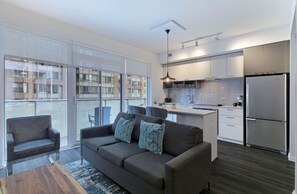 A modern apartment with a grey sofa, a dining area, a kitchen with white cabinets, and a large window overlooking a building. - 3 Bedroom/2 Bathroom Suite (Toronto)