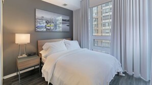 Hotellihuone, jossa on sänky, yöpöytä lampun kanssa, suuri ikkuna verhoilla ja kaupunkimaisemaa esittävä taulu seinällä.