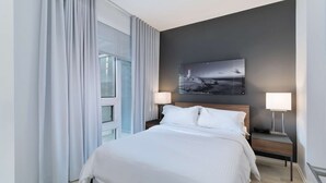 Une chambre d’hôtel avec un grand lit, deux tables de chevet avec des lampes, une table d’appoint, une fenêtre avec des rideaux et une œuvre d’art accrochée au mur.