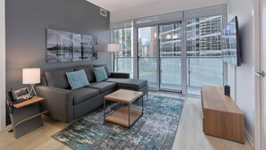 Un salon moderne avec un canapé gris, une table basse, un téléviseur et une vue sur la ville à travers de grandes fenêtres.
