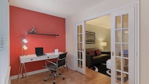 Une pièce avec un mur rouge, un bureau blanc avec un ordinateur portable, une lampe sur pied et une chaise. Il y a une porte qui donne sur une autre pièce, où se trouvent un canapé et une lampe.