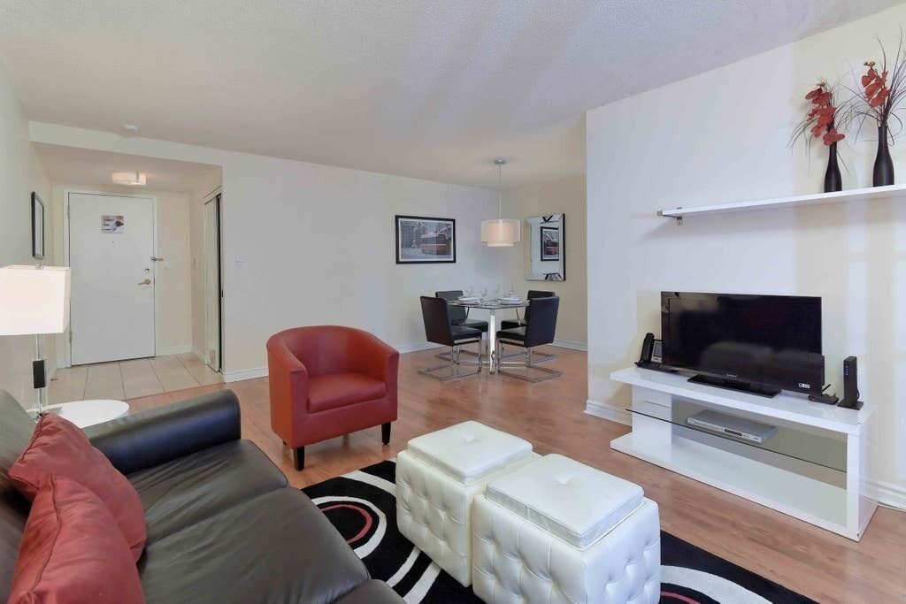 Una sala de estar moderna con un sofá, otomanas, una silla roja, una mesa de comedor con sillas, un televisor sobre un mueble blanco y una pequeña estantería con objetos decorativos.