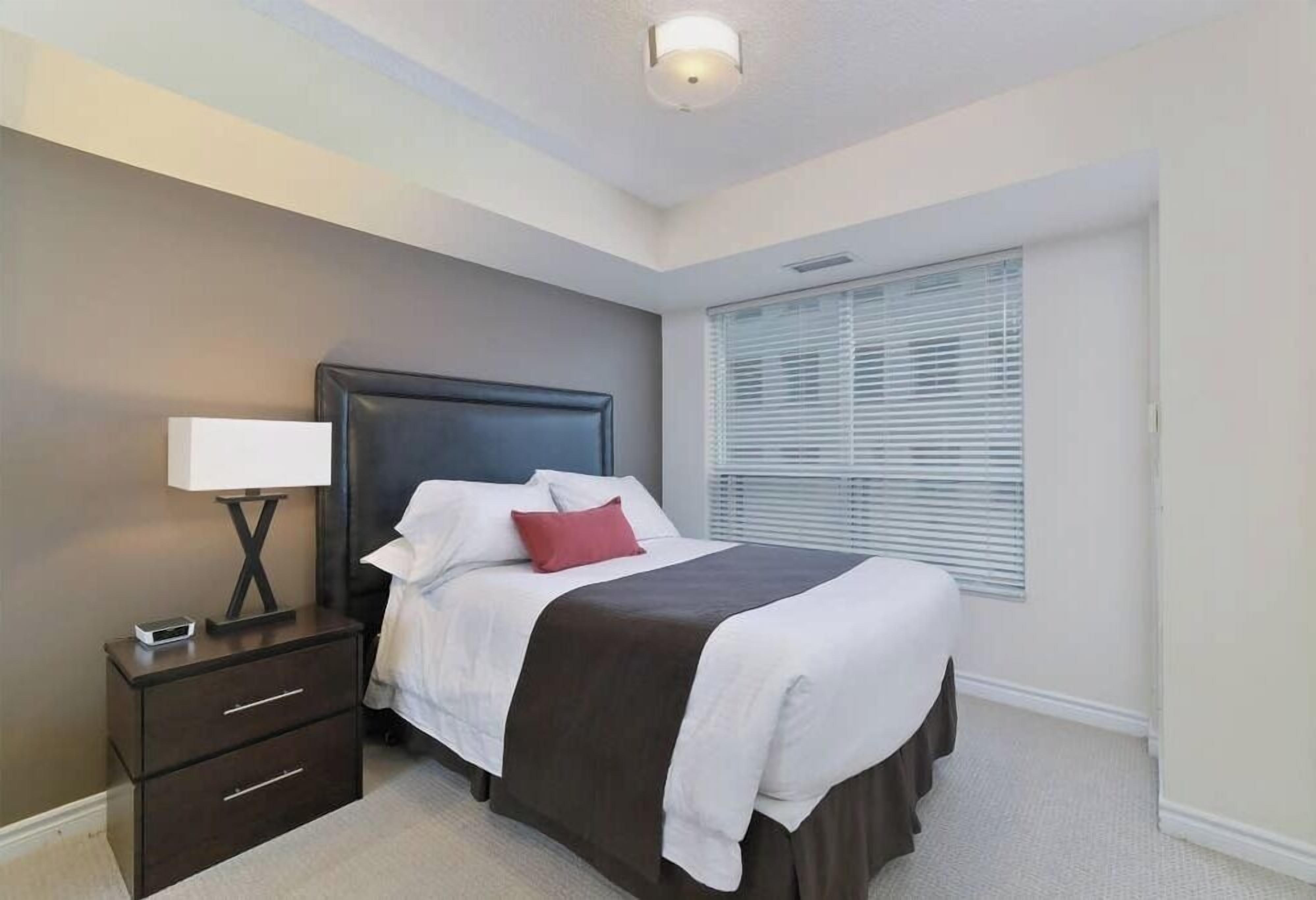 A bedroom with a dark headboard, a white bedspread, a red pillow, a bedside table with a lamp, and a window with blinds.
