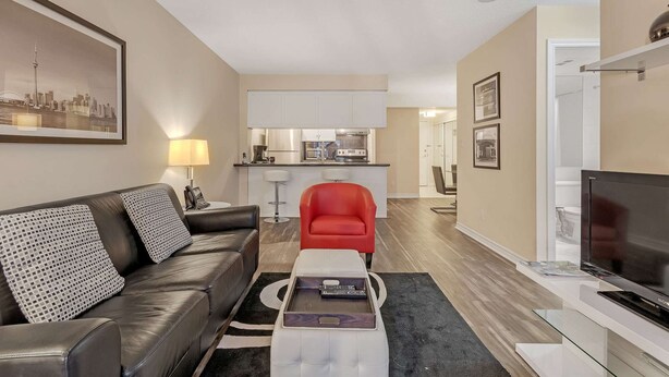 Un salon avec un canapé, un fauteuil rouge, un téléviseur et, au fond, une cuisine.