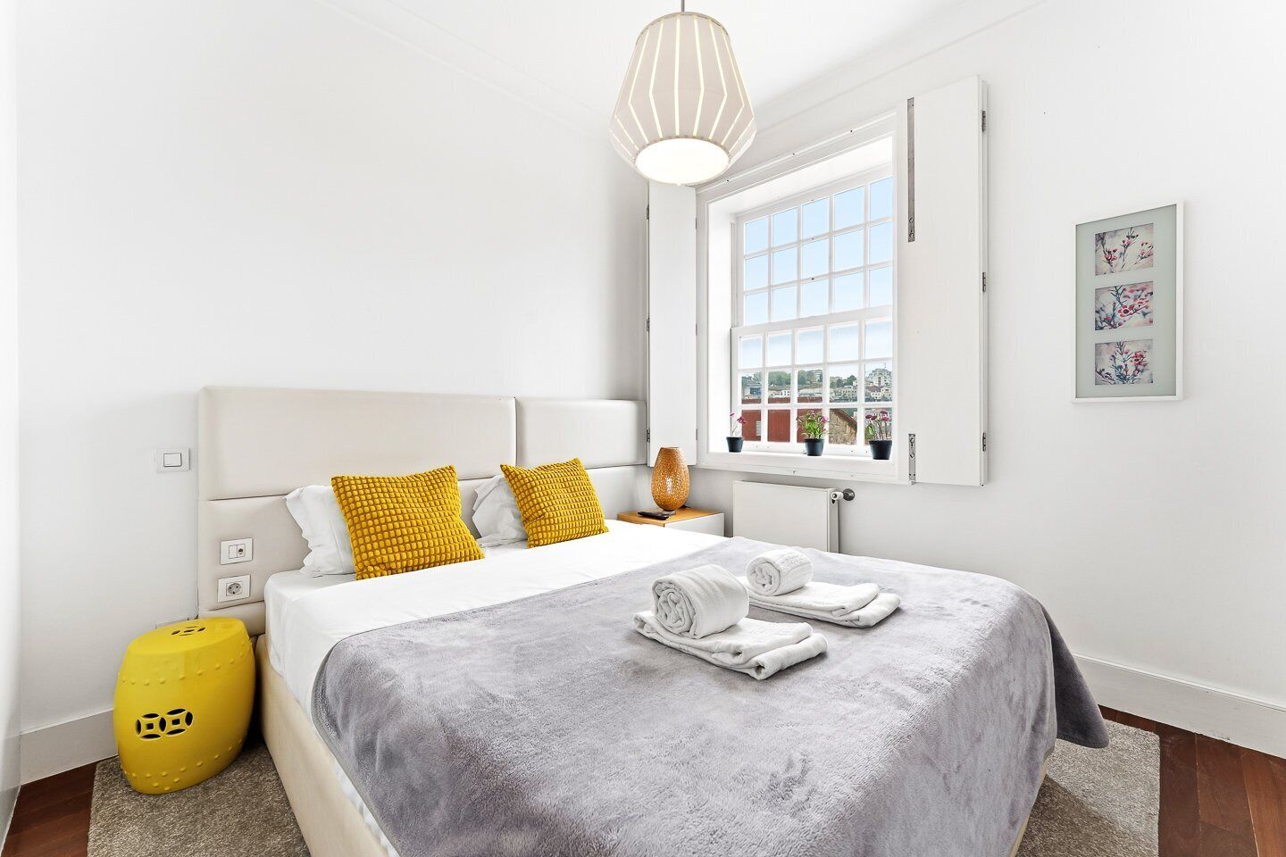 A modern hotel room with a bed, a window with a view, a bedside table, and a yellow ottoman.