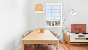 A modern room with a wooden dining table, chairs, a lamp, a TV, and a window with a city view. - GuestReady - Ribeira retreat in Porto centre (Porto)