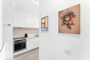 A modern kitchen with white cabinets, a coffee machine, and a microwave. There is a framed pasta chart and a painting of apples on the wall.