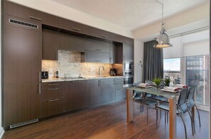 A modern kitchen with dark wood cabinets, a granite countertop, and stainless steel appliances. There is a dining area with a glass table and chairs, and a view of the city through large windows. - 1 Bedroom/1 Bathroom Suite (Toronto)