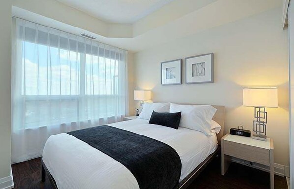 A hotel room with a large bed, two bedside lamps, a nightstand with a radio, and a window with sheer curtains. - 2 Bedroom/2 Bathroom Suite (Toronto)