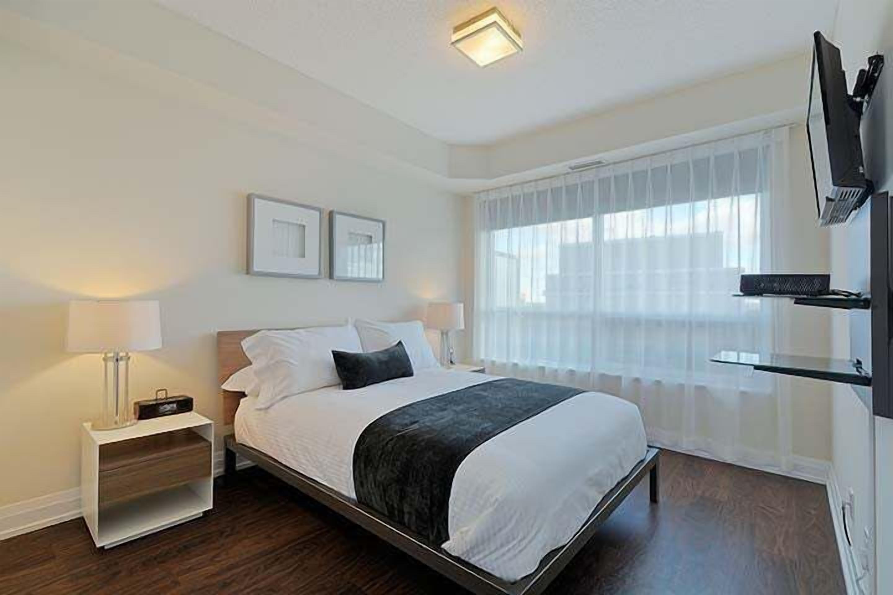 Une chambre d’hôtel moderne avec un grand lit, une table de chevet avec une lampe, un téléviseur fixé au mur et une fenêtre avec des rideaux légers.