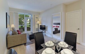 A modern hotel room with a dining area, a sofa, a TV, and a bed with a view of the outdoors.