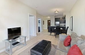 A modern living room with a flat-screen TV on a glass stand, a grey sofa with red cushions, and a black ottoman.