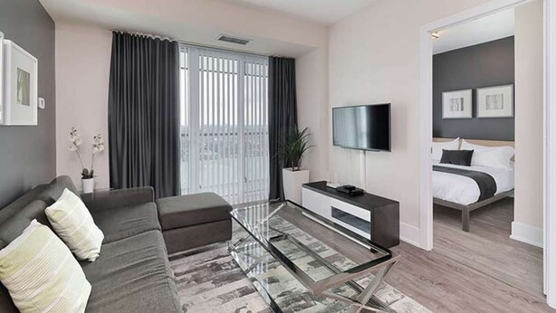 A modern living room with a sofa, coffee table, and a TV. There is a balcony with curtains and a bed in the adjacent room. - 2 Bedroom/2 Bathroom Suite (Toronto)