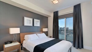 Une chambre d’hôtel avec un grand lit, deux tables de chevet, une vue sur la ville et deux tableaux encadrés au mur.