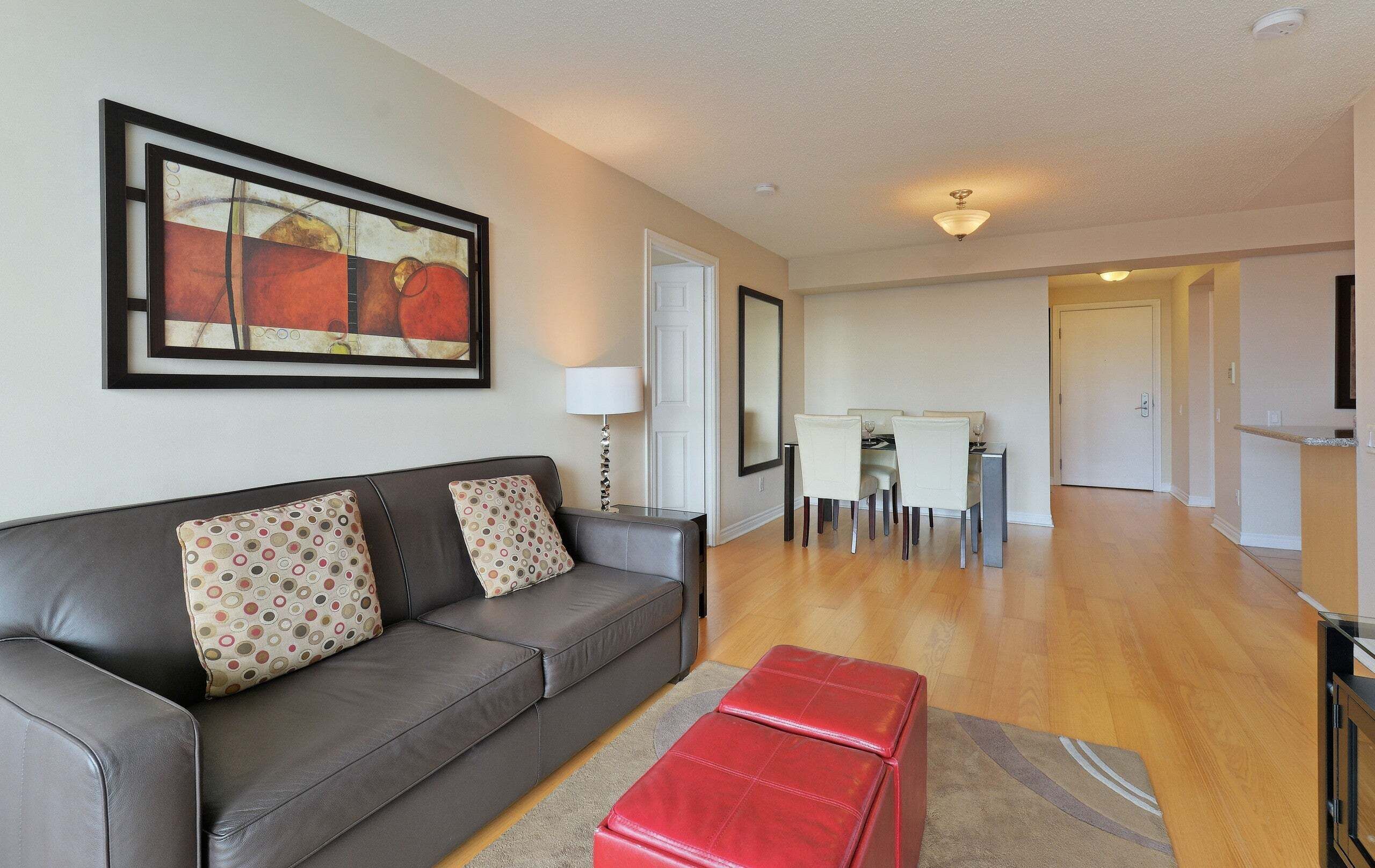 A modern living room with a grey sofa, red ottoman, and a framed abstract painting.