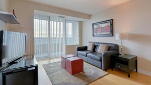 A modern living room with a flat-screen TV, a red ottoman, a grey sofa with patterned cushions, a side table with a lamp, and a large window with blinds. - 2 Bedroom/2 Bathroom Suite (Scarborough ON)