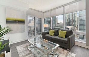 A modern living room with a gray sofa, a glass coffee table, and large windows offering city views.