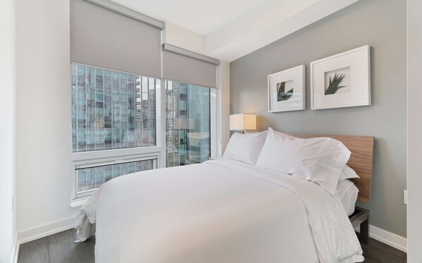 A hotel room with a large window, a bed with white linens, a nightstand with a lamp, and two framed pictures on the wall.