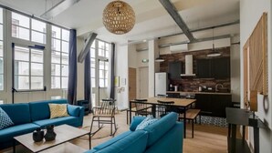 A modern living room with a blue sofa, a dining table, and a kitchen area with a brick wall and wooden cabinets. - PHILIPPE AUGUSTE LOFT 2 - Loft for 10 - 4 rooms - BASTILLE - PÈRE LACHAISE (Paris)