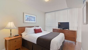 Hotelkamer met een bed, nachtkastje met lamp, tv op een houten dressoir en een raam met zonwering.