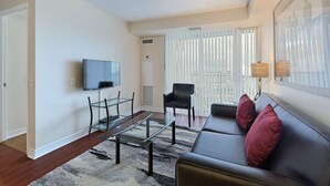 Een moderne woonkamer met een flatscreen televisie, een glazen salontafel, een leren bank met rode kussens en een groot raam met jaloezieën.