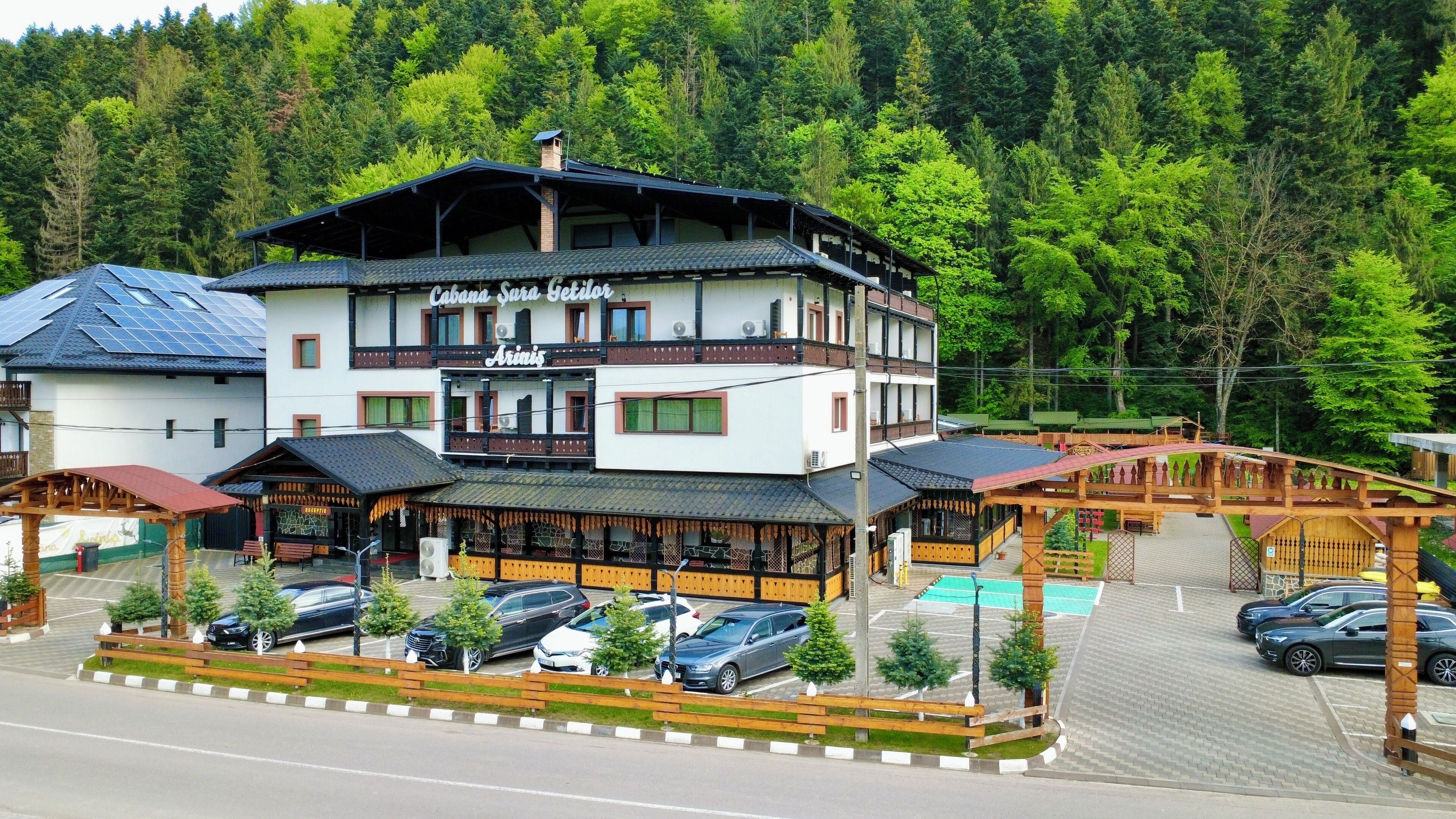 A multi-story building with a restaurant named "Cabana Sura Sella" and "Arinis" on the ground floor. There are several cars parked in front, and a wooden bridge connects different parts of the property.