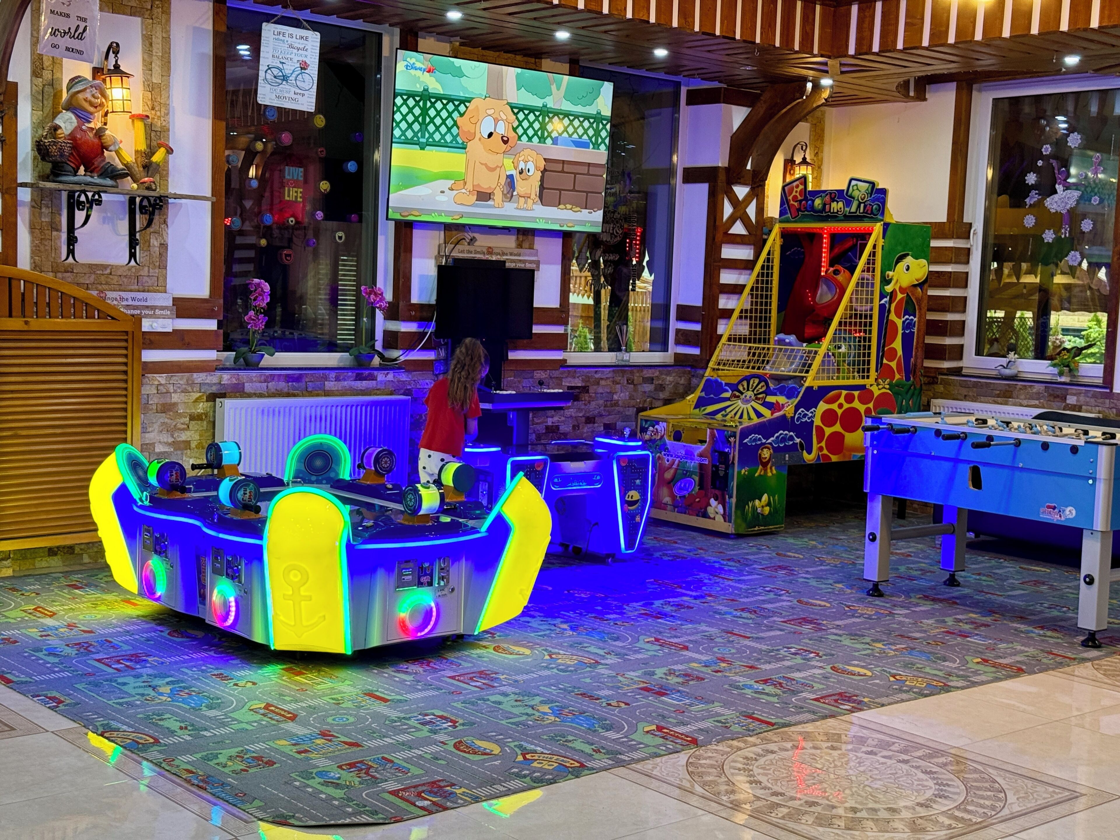 A brightly lit indoor play area with a foosball table, arcade machines, and a television screen displaying a cartoon.
