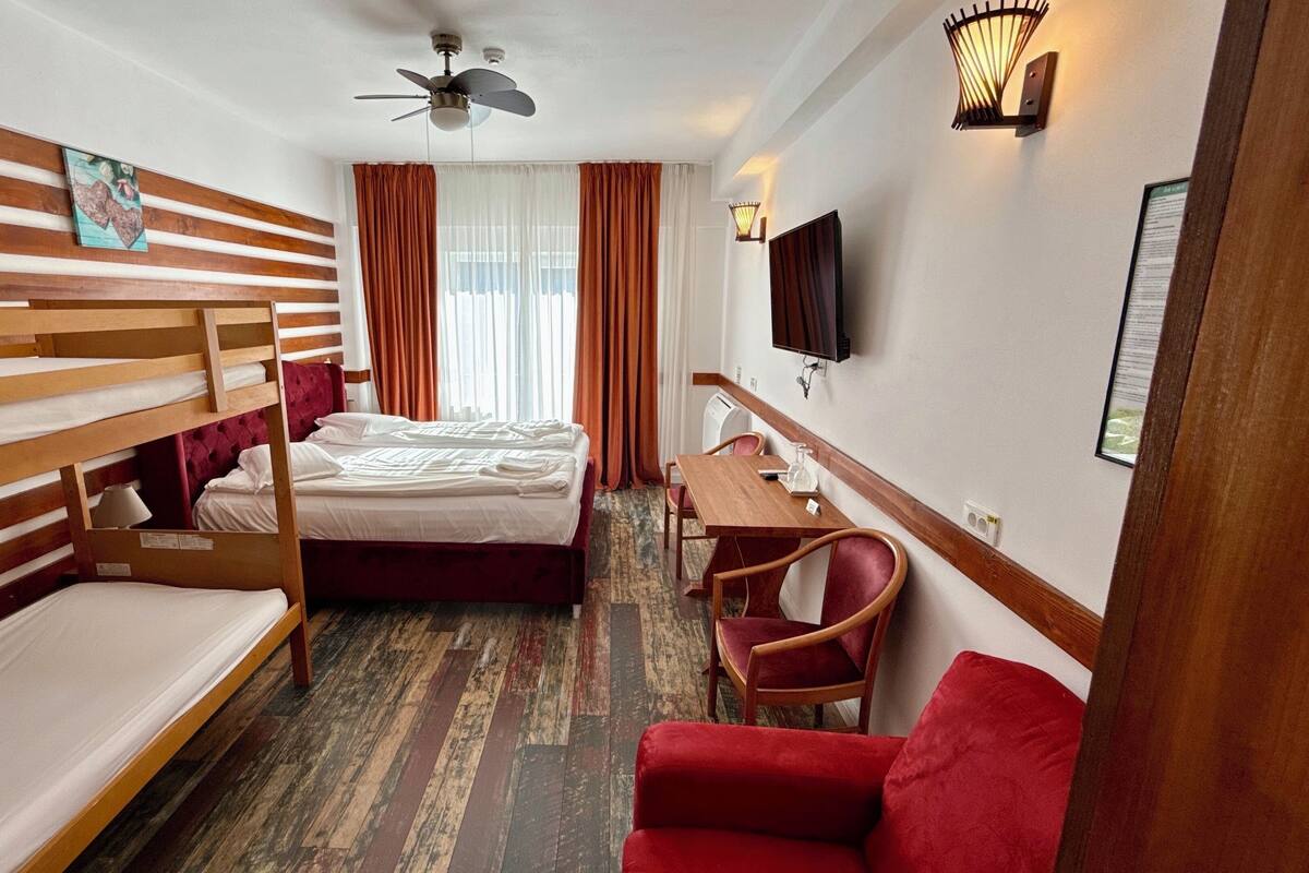 Een hotelkamer met een stapelbed, een eenpersoonsbed, een bureau met stoel, een televisie en een rode bank.