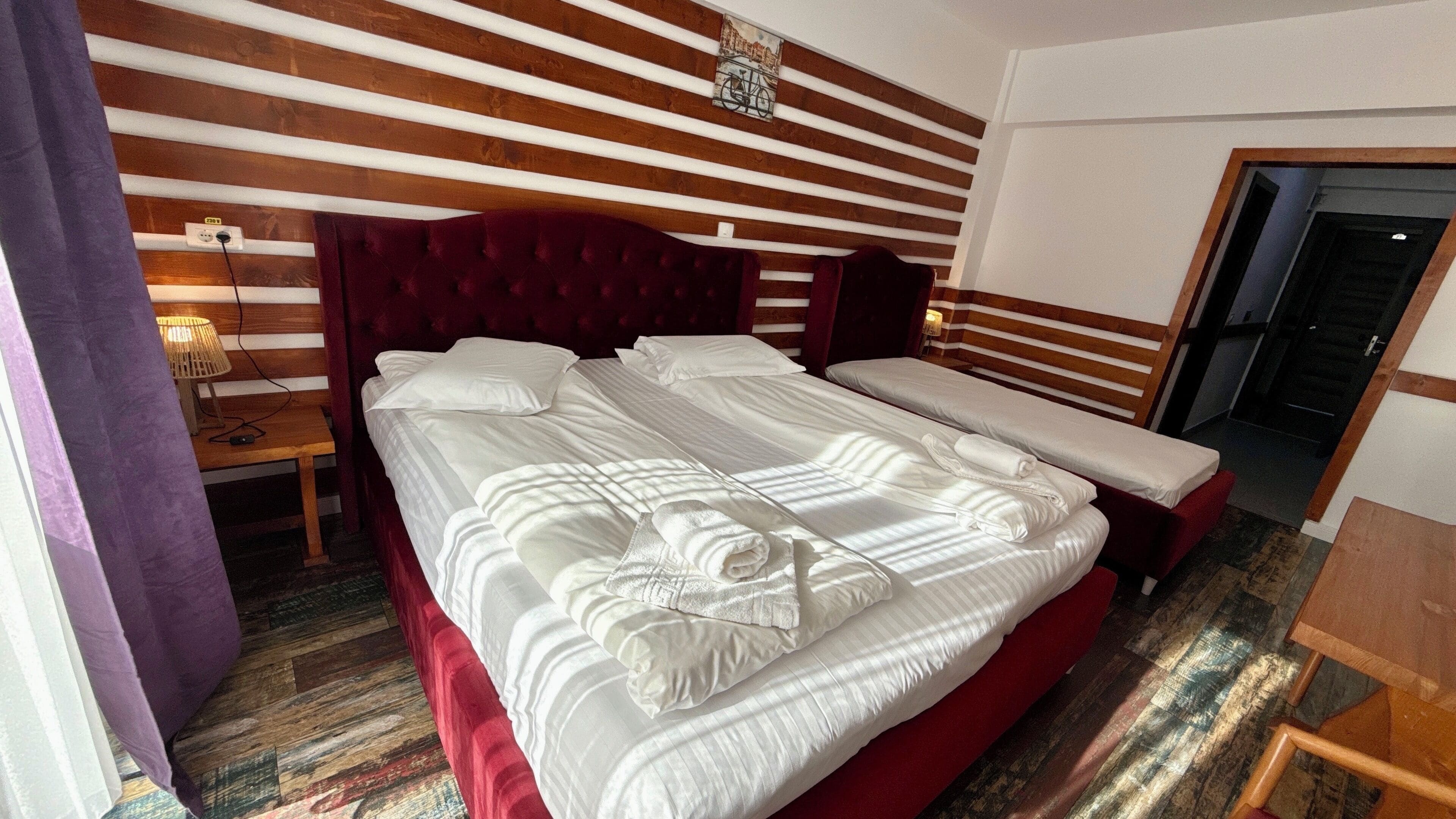 A hotel room with a large bed, a smaller bed, a wooden headboard, a nightstand with a lamp, and a wooden floor.