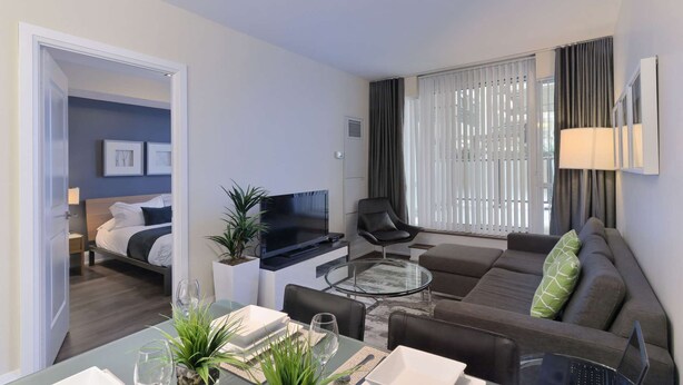 Ein modernes Hotelzimmer mit Bett, Fernseher, Sofa, Essbereich und einem großen Fenster mit Vorhängen.