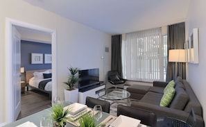 Ein modernes Hotelzimmer mit Bett, Fernseher, Sofa, Essbereich und einem großen Fenster mit Vorhängen.