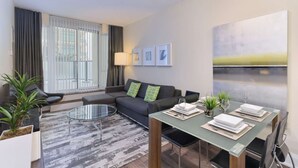 Ein modernes Wohnzimmer mit einer Couch, einem Glastisch, einem Esstisch für zwei Personen und einem großen Fenster mit Vorhängen.
