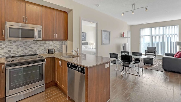 A modern kitchen with stainless steel appliances, wooden cabinets, and a large island. There is a dining area with a table and chairs, and a living room with a sofa and a large window.