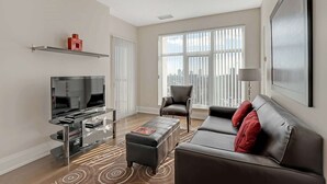 A modern living room with a flat-screen TV, a leather sofa, a coffee table, and a large window offering a city view.