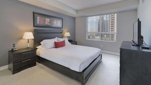 Ein modernes Hotelzimmer mit einem großen Bett, zwei Nachttischen mit Lampen, einem Schminktisch, einem Fernseher und einem Fenster mit Jalousien.