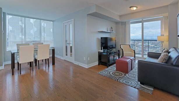 A spacious living area with a dining table, chairs, a sofa, a TV, and a large window offering a city view. - 2 Bedroom/2 Bathroom Suite (Etobicoke)