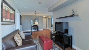 A living room with a brown leather sofa, a red ottoman, a flat-screen TV on a black stand, and a patterned rug. - 2 Bedroom/2 Bathroom Suite (Etobicoke)