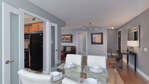 A modern dining area with a glass table set for four, white chairs, and a kitchen visible through an open door. - 2 Bedroom/2 Bathroom Suite (Etobicoke)