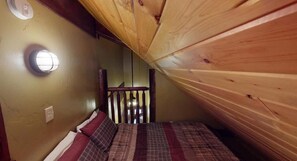 A small, cozy room with a sloped ceiling, a bed with plaid bedding, a round wall light, and a wooden railing.