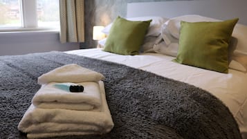 Una cama bien tendida con una manta gris con textura, dos almohadas verdes y una toalla blanca doblada con un objeto negro encima.