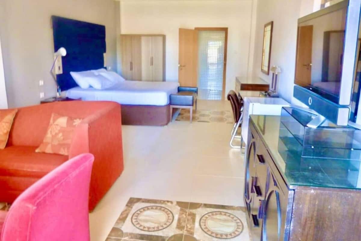 A hotel room with a bed, a red sofa, a TV, and a desk with a mirror.