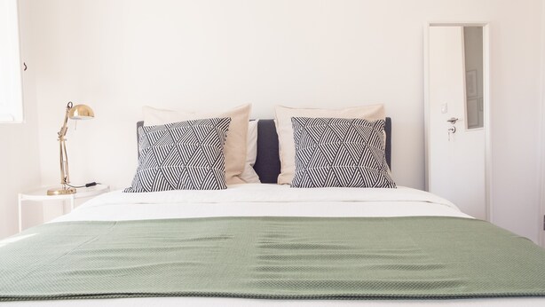 A neatly made bed with a green blanket, two patterned pillows, and a bedside lamp on a white nightstand. - Bairro Conqvistador Apartment 4 (Lisboa)
