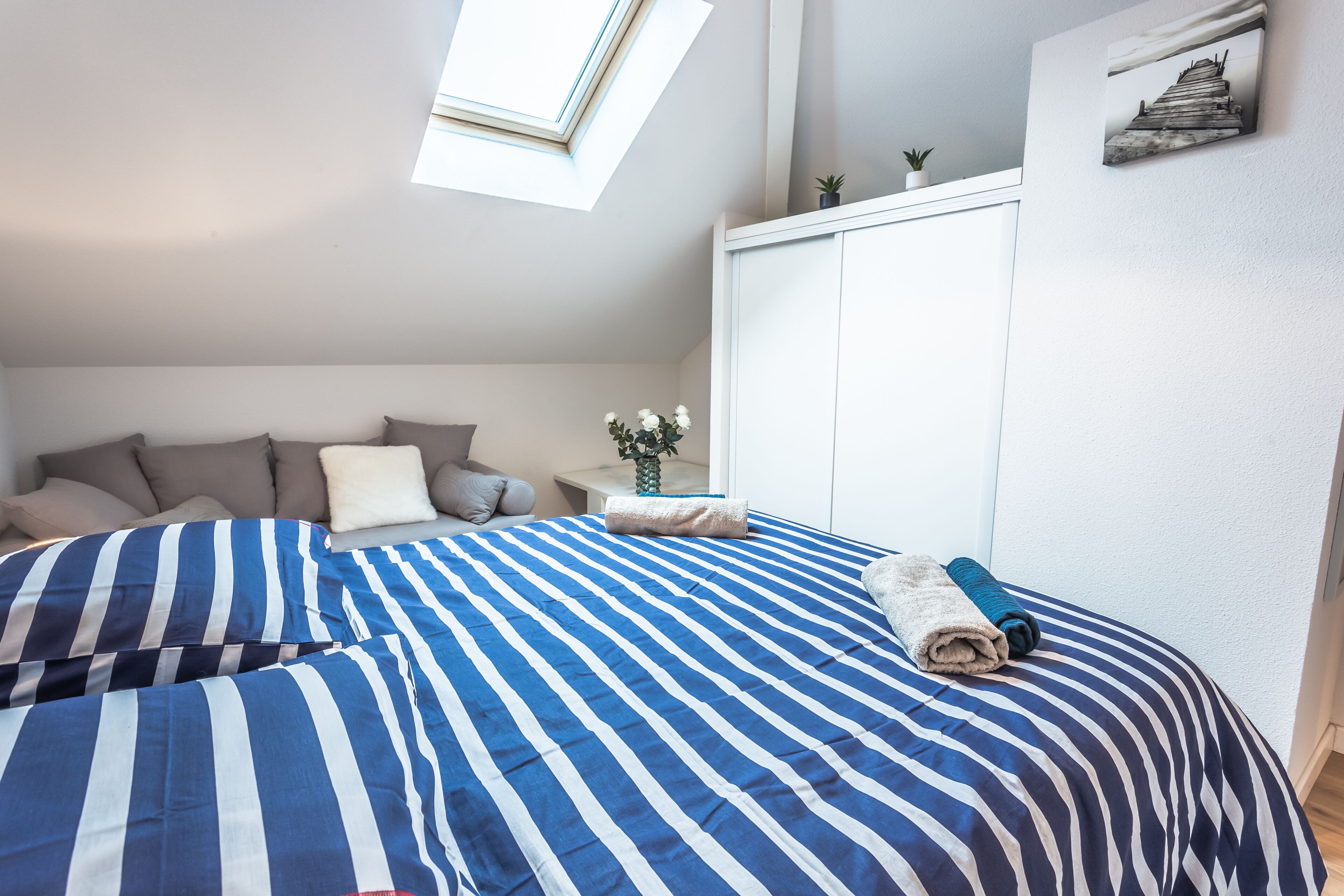 Ein Schlafzimmer mit Dachfenster, gestreifter Bettwäsche, einem Bett mit Kissen, einem Nachttisch mit Pflanze und einem Kleiderschrank.