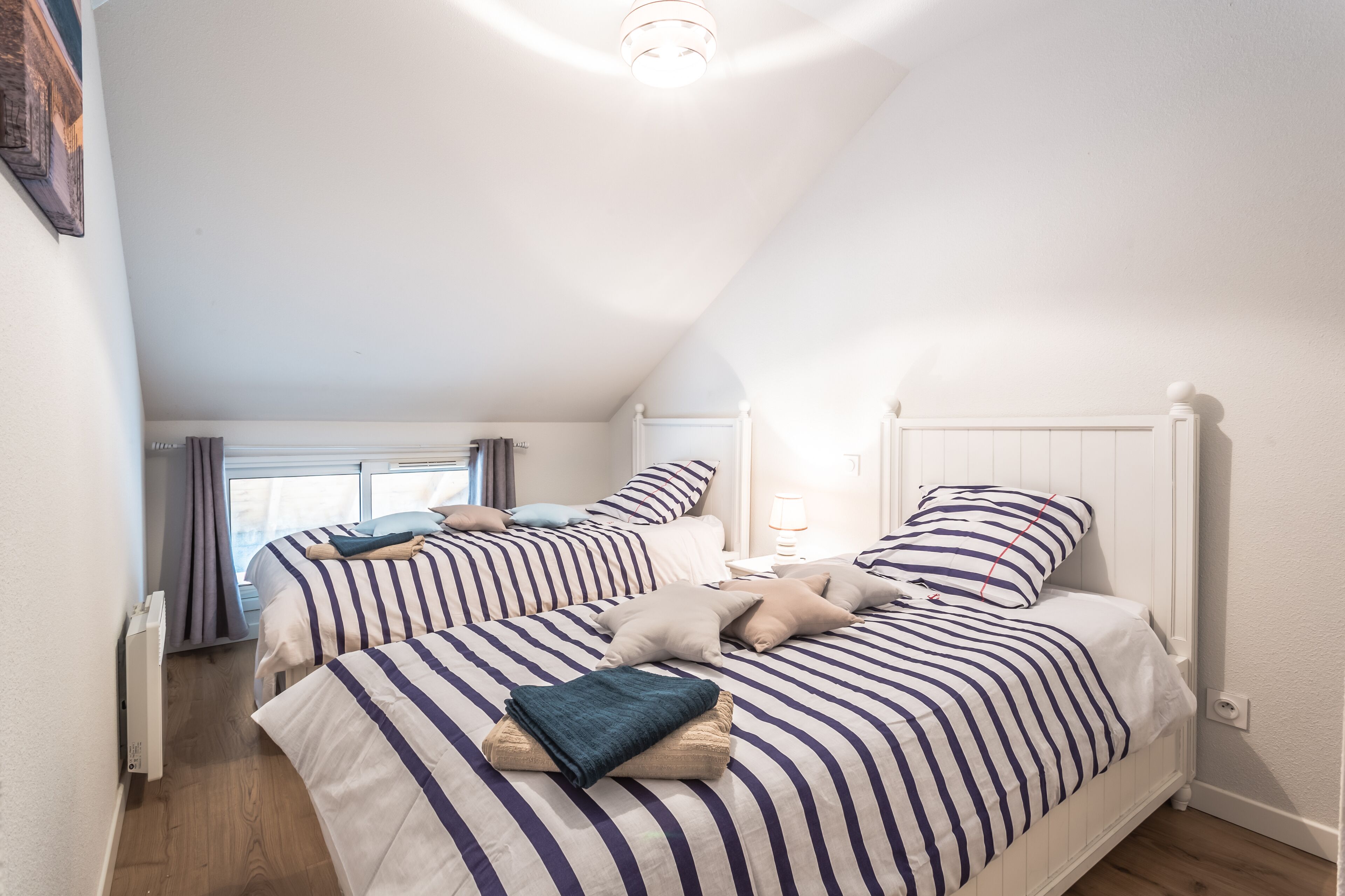 A bedroom with two beds, each with striped bedding and pillows, a bedside table with a lamp, and a window with curtains.