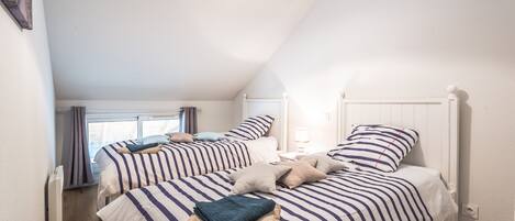 A bedroom with two beds, each with striped bedding and pillows, a bedside table with a lamp, and a window with curtains.