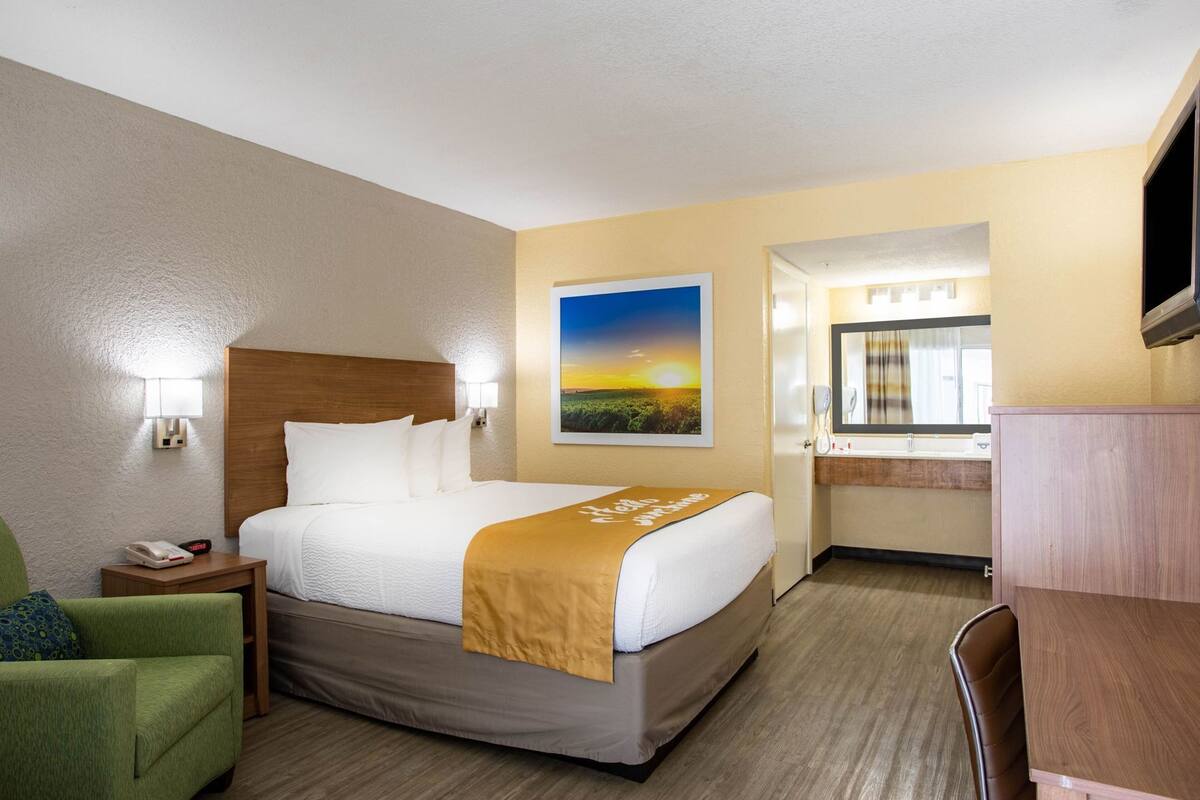 A hotel room with a bed, bedside table, lamp, chair, and a painting of a sunset.